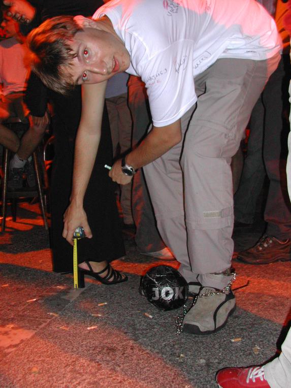 A man in a white shirt bends down while wearing a ball and chain, as someone measures the distance.
