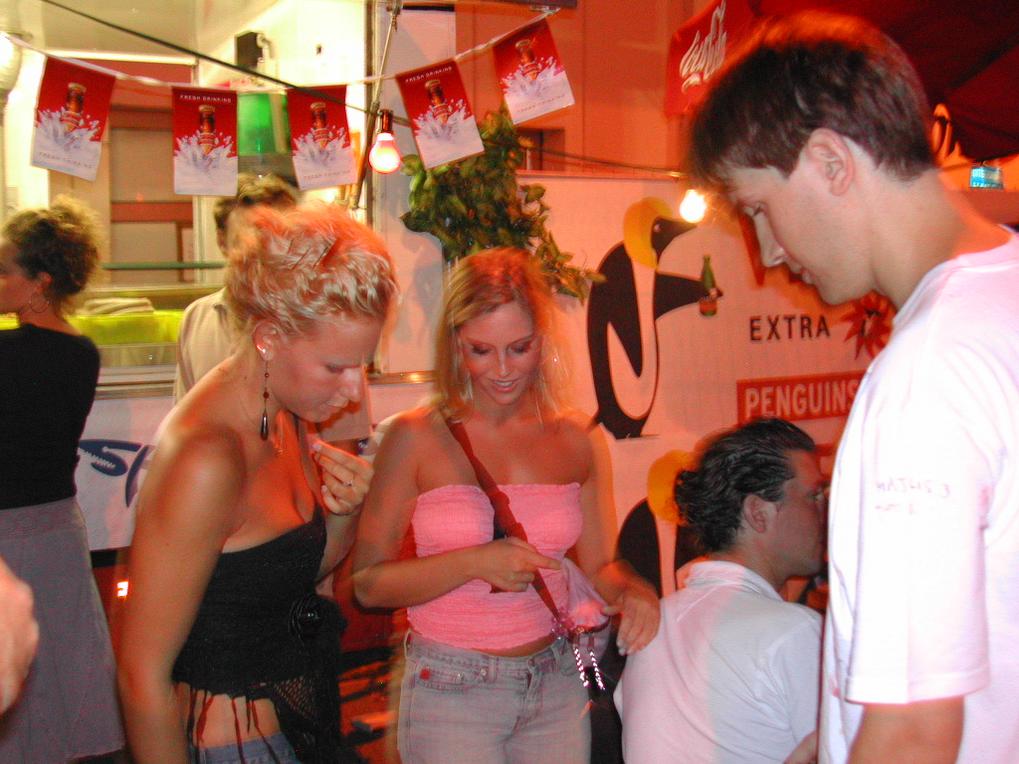 A group of people socializing at a bar, with two women examining an item in a lively setting.