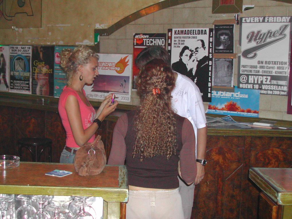 A woman in a pink top talks to two people at a bar, holding a small object in her hand.