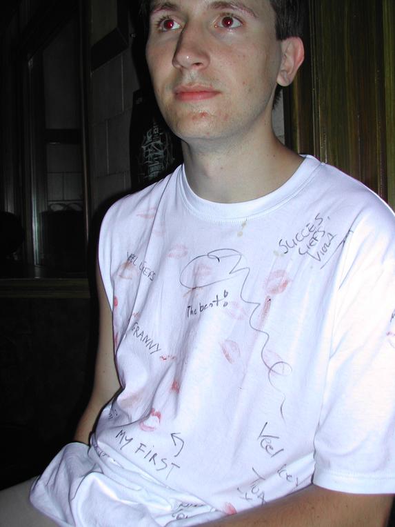 A man wearing a white T-shirt covered in lipstick marks and handwritten messages sits in a dimly lit bar.