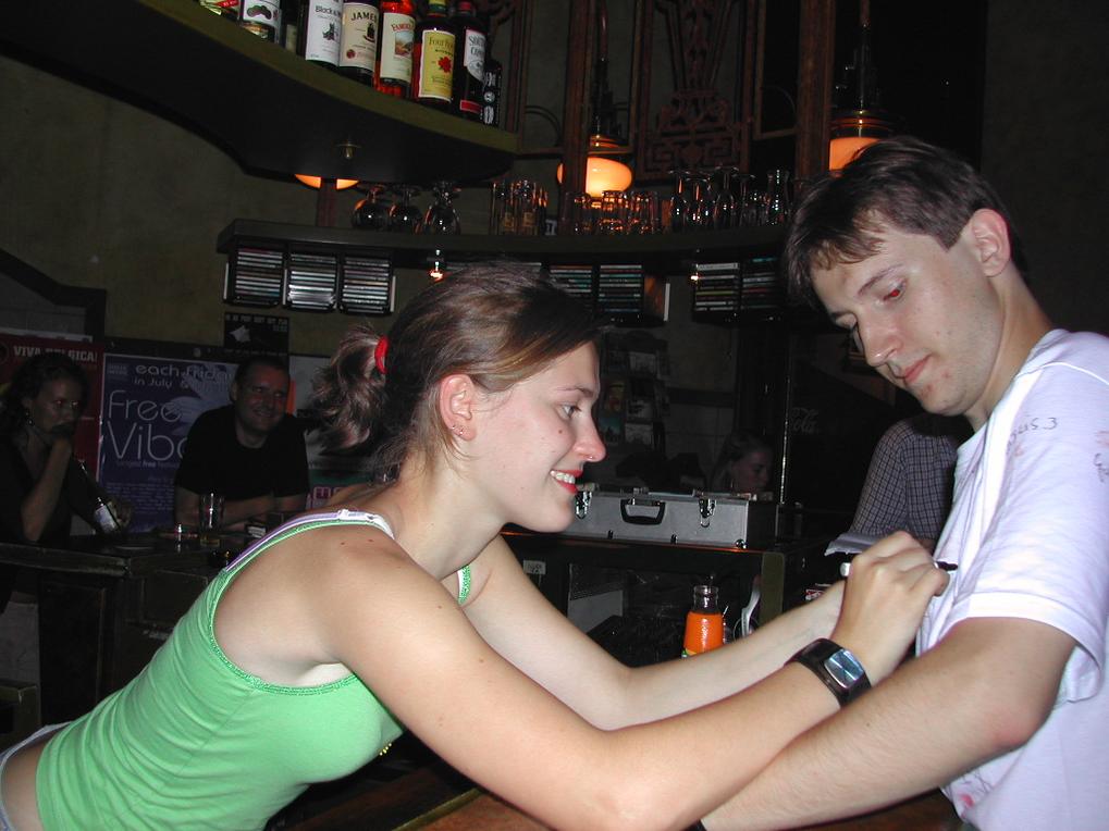 A woman in a green top writes on a man's white shirt at a bar, both smiling.