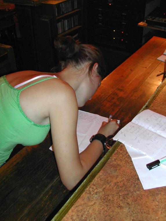 A woman in a green top leans over a bar counter, writing on a piece of paper with a pen.