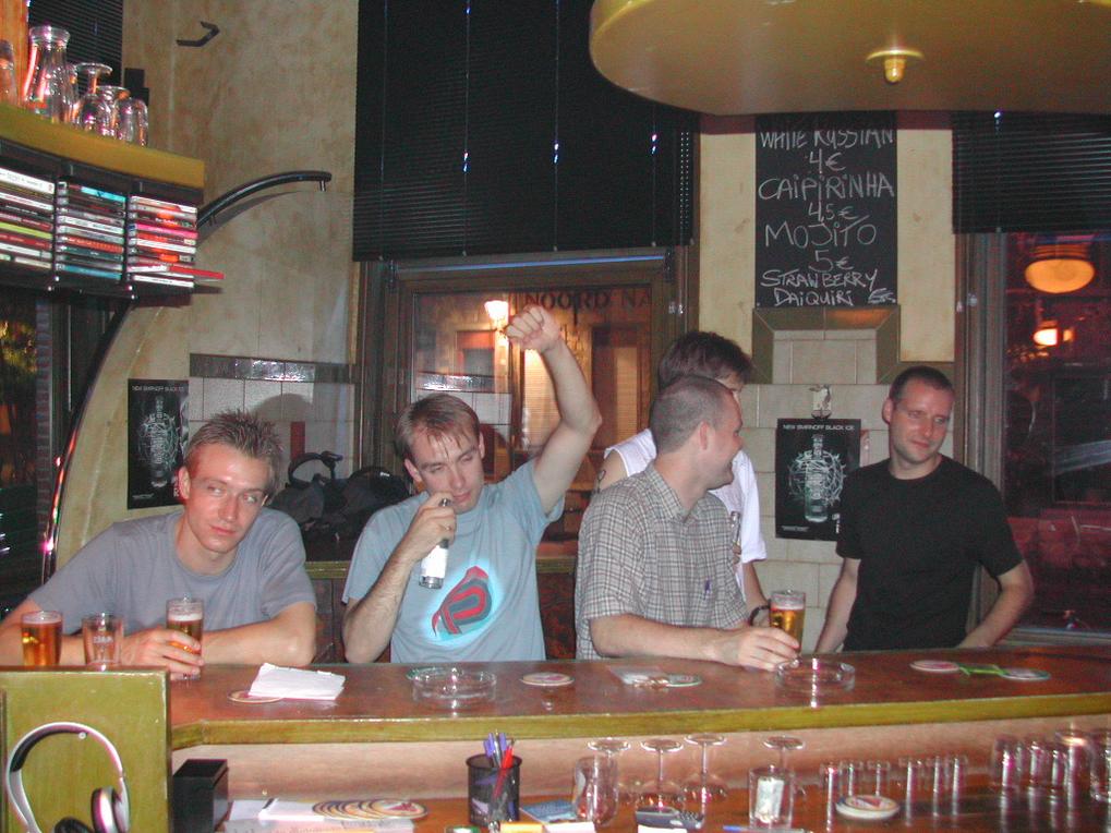 A group of men at a bar, one raising his arm while holding a drink, others engaged in conversation.