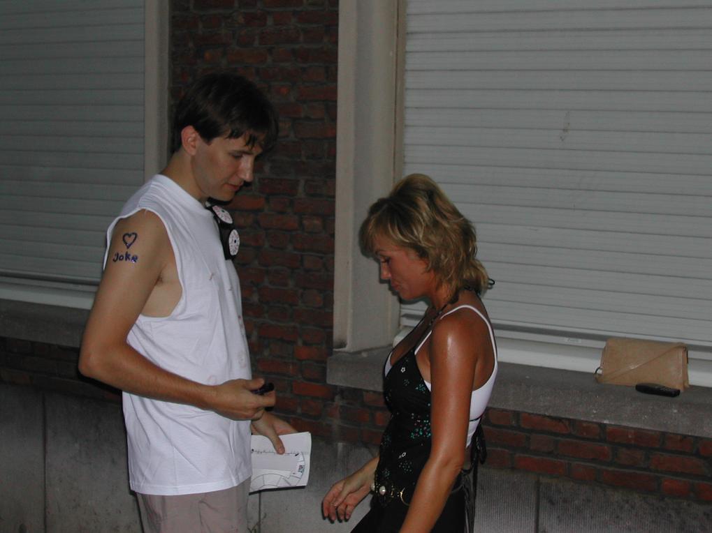 A man in a white shirt with writing on his arm looks at a woman in a black outfit outside.