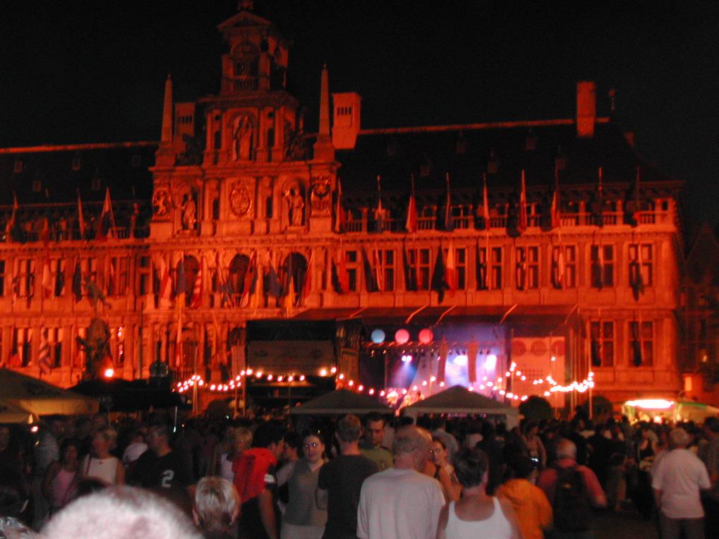 A large crowd gathers at night in front of an illuminated historic building with a stage and festive lights.
