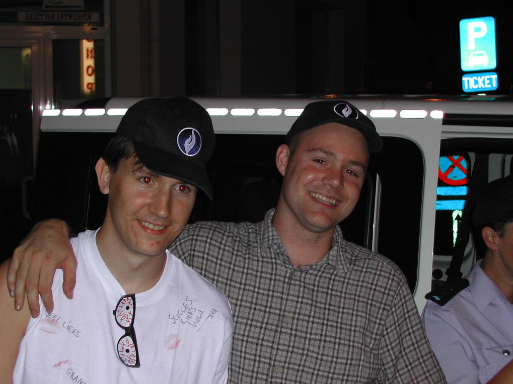 Two men smiling and posing together at night, both wearing matching black hats with a logo.