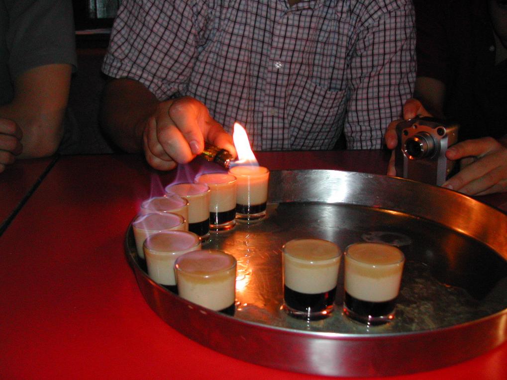 A person lights shots on fire while another takes a photo at a bar.