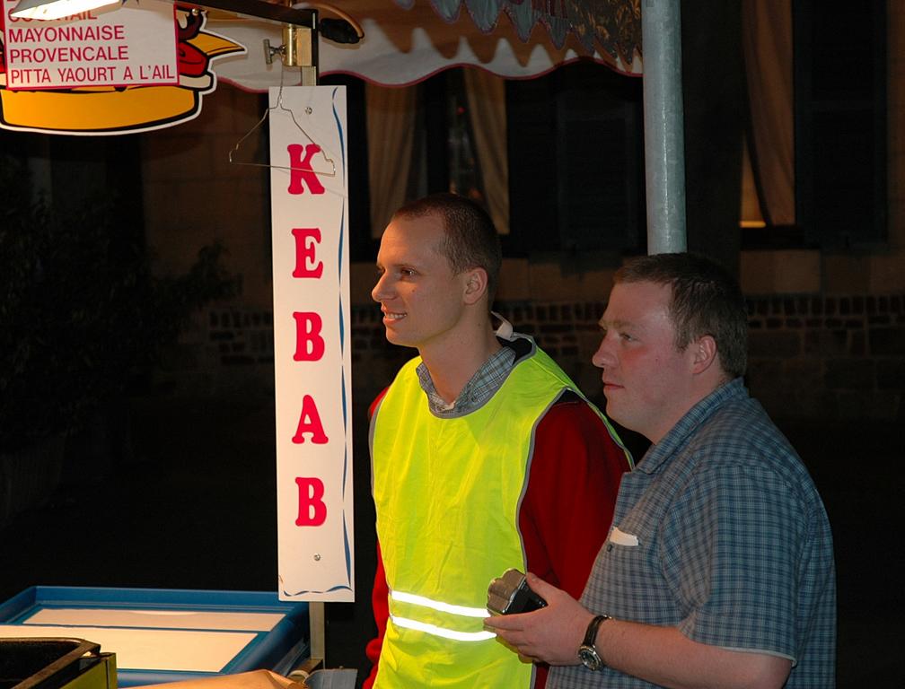 Two men stand near a kebab stand at night, one wearing a reflective vest and the other holding something.