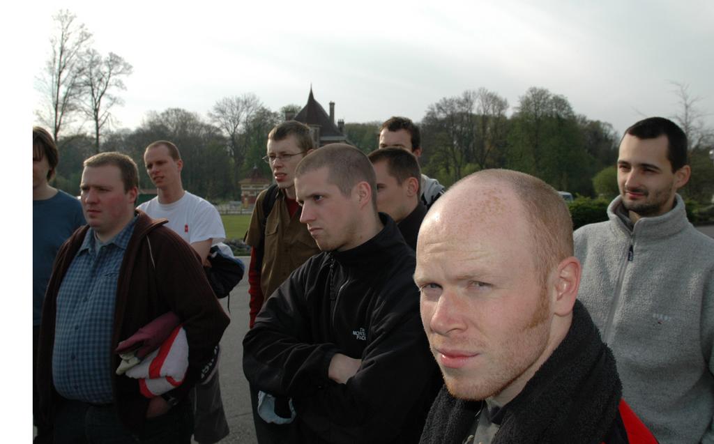 A group of men standing outdoors, with one bald man in the foreground looking directly at the camera.