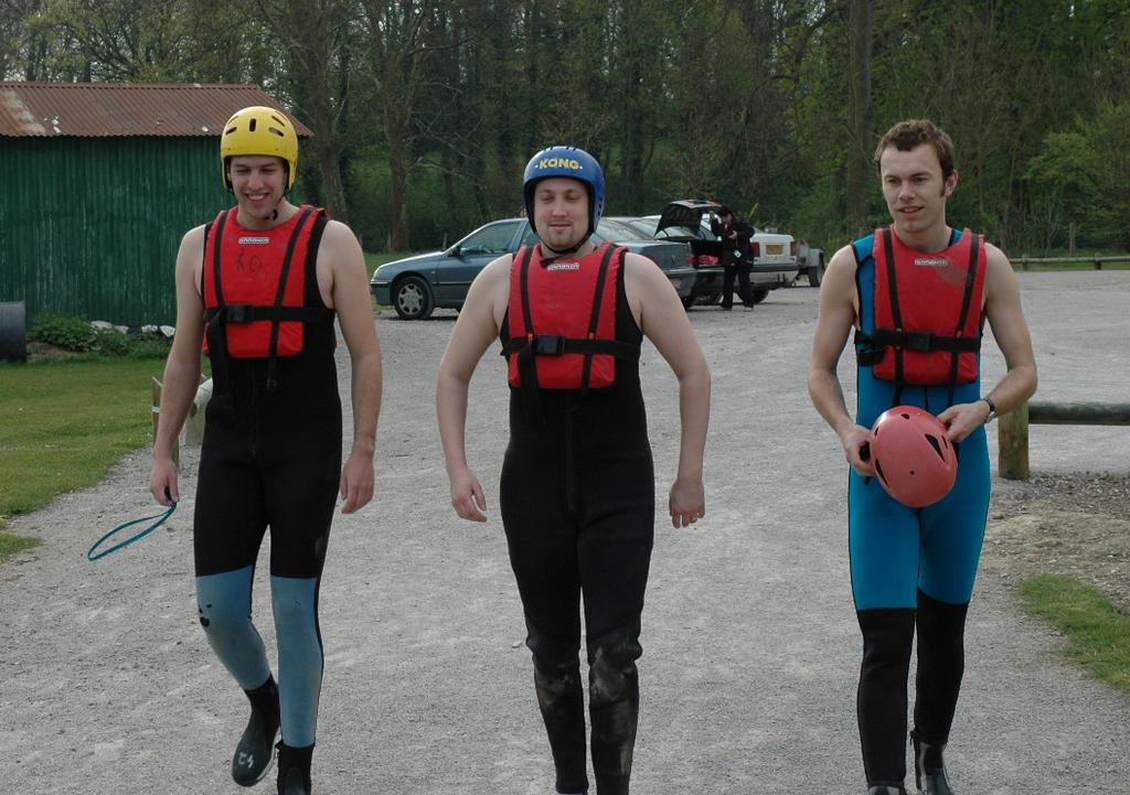 Three men in wetsuits and life jackets walk on a gravel path, one holding a helmet.