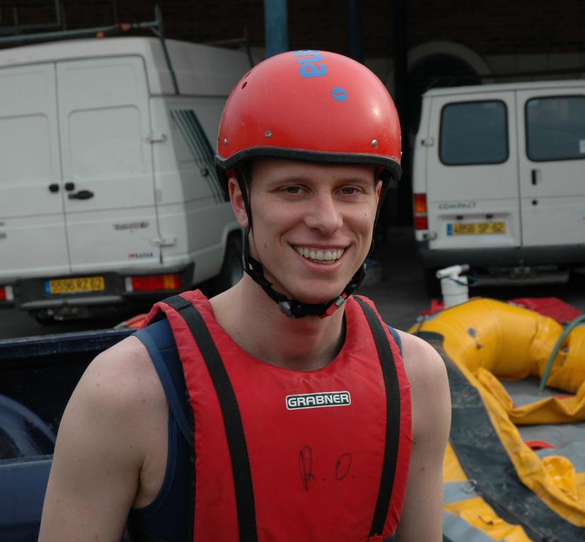 A man wearing a red helmet and life vest smiles at the camera, standing near inflatable rafting equipment.