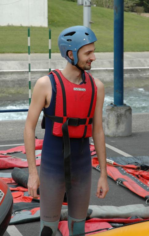 A man wearing a helmet, life jacket, and wetsuit stands near rafting equipment, looking to the side.