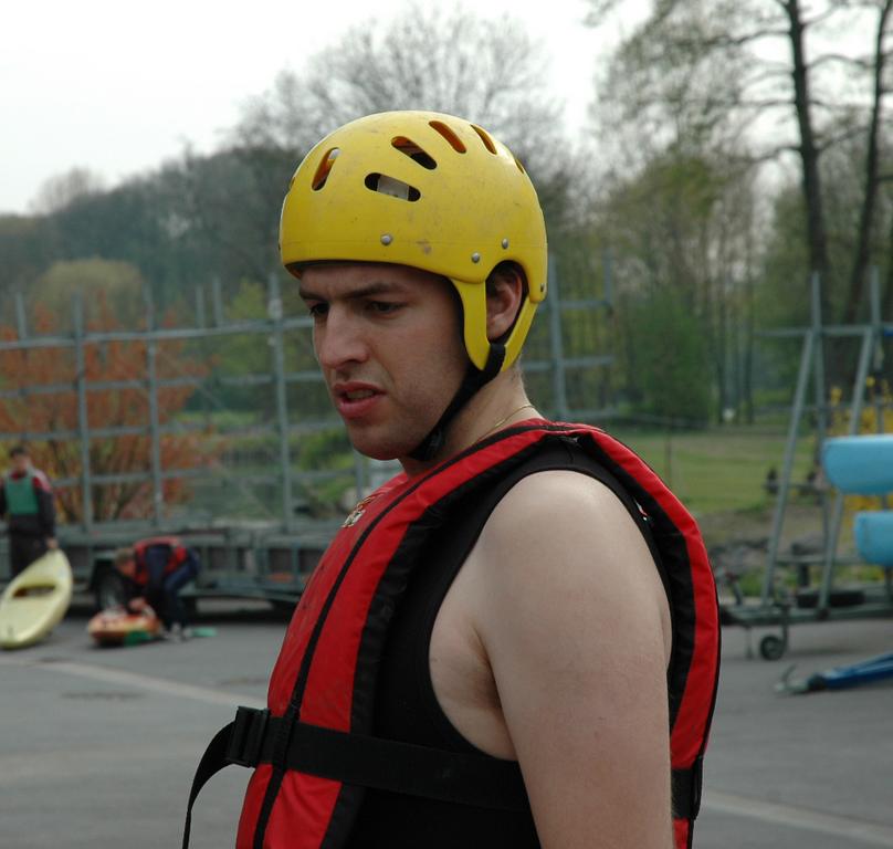 A man wearing a yellow helmet and red life jacket stands outdoors, looking to the side with a focused expression.