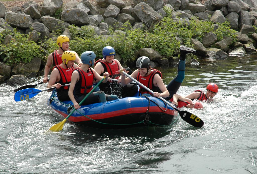 A group of people in helmets and life jackets raft on a river, while one person falls into the water.