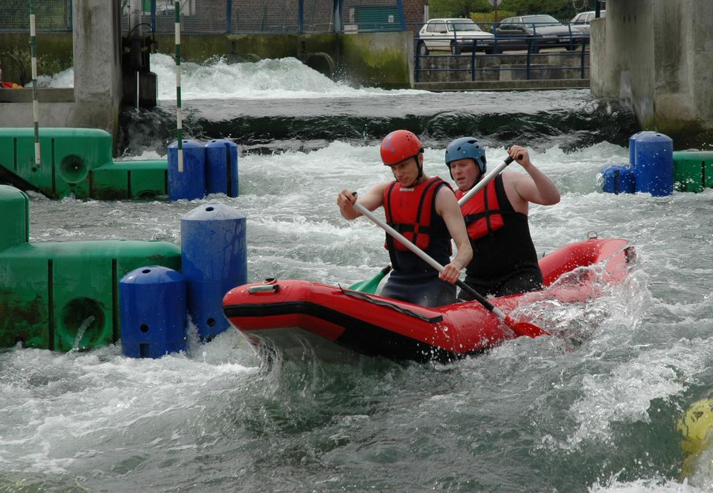 Two people in helmets and life jackets paddle a red inflatable raft through whitewater rapids.
