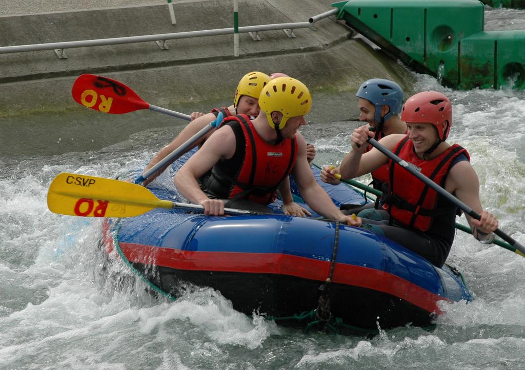 Four men in helmets and life jackets paddle a blue inflatable raft through whitewater rapids, smiling and enjoying the ride.