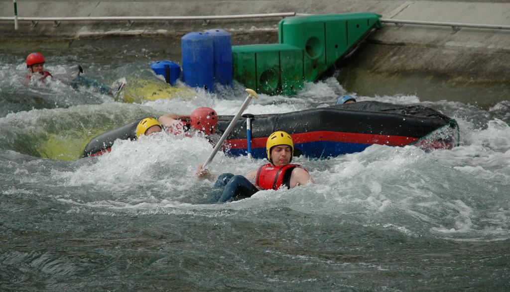A man in a yellow helmet and red vest holds a paddle while floating in rough water near an overturned raft.