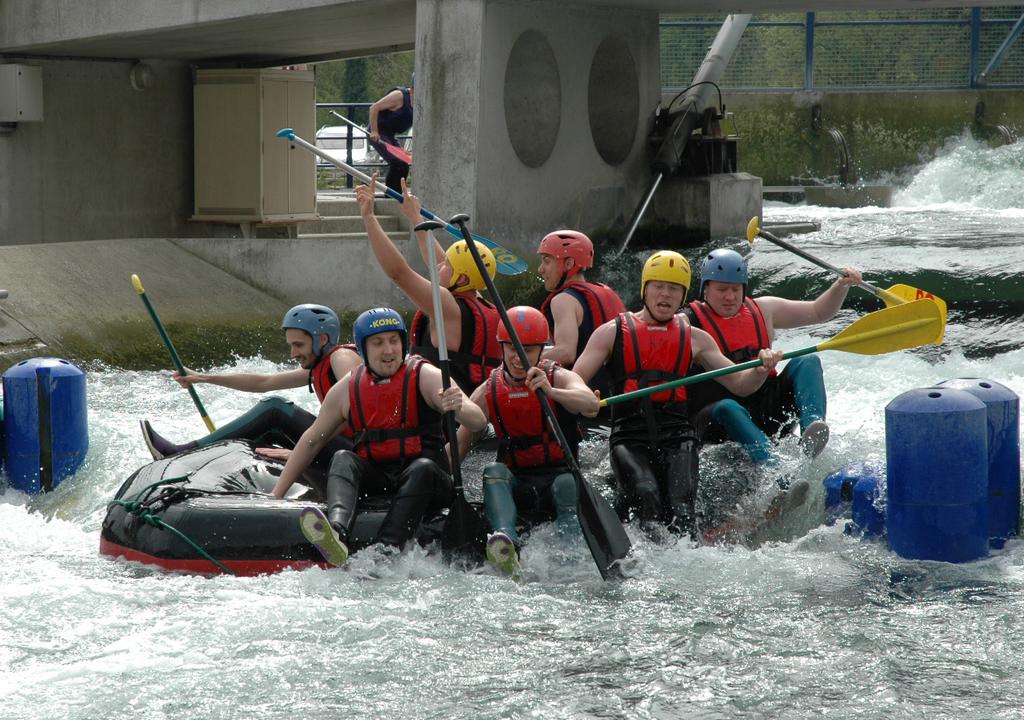 A group of people wearing helmets and life jackets paddle a raft through rough water, appearing excited and focused.