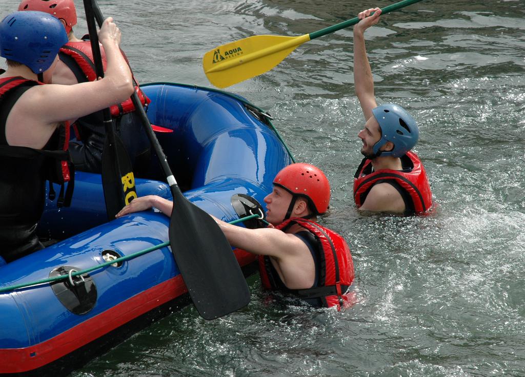 Two people in helmets and life jackets are in the water, reaching for a raft while others assist them.