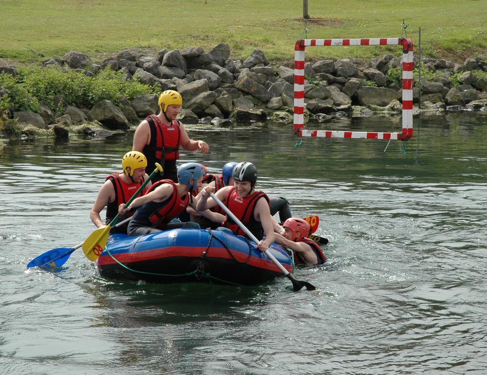 A group of people in helmets and life jackets paddle a raft while one person struggles to climb aboard.