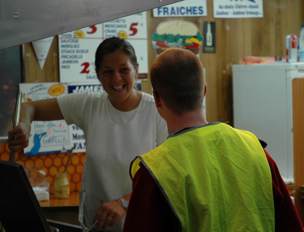 A woman behind a food counter smiles while talking to a man wearing a yellow vest.