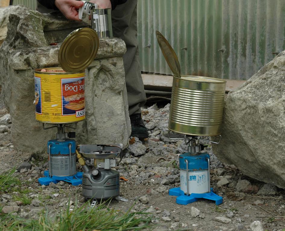 A person opens a can while two makeshift stoves heat food in large cans outdoors on rocky ground.