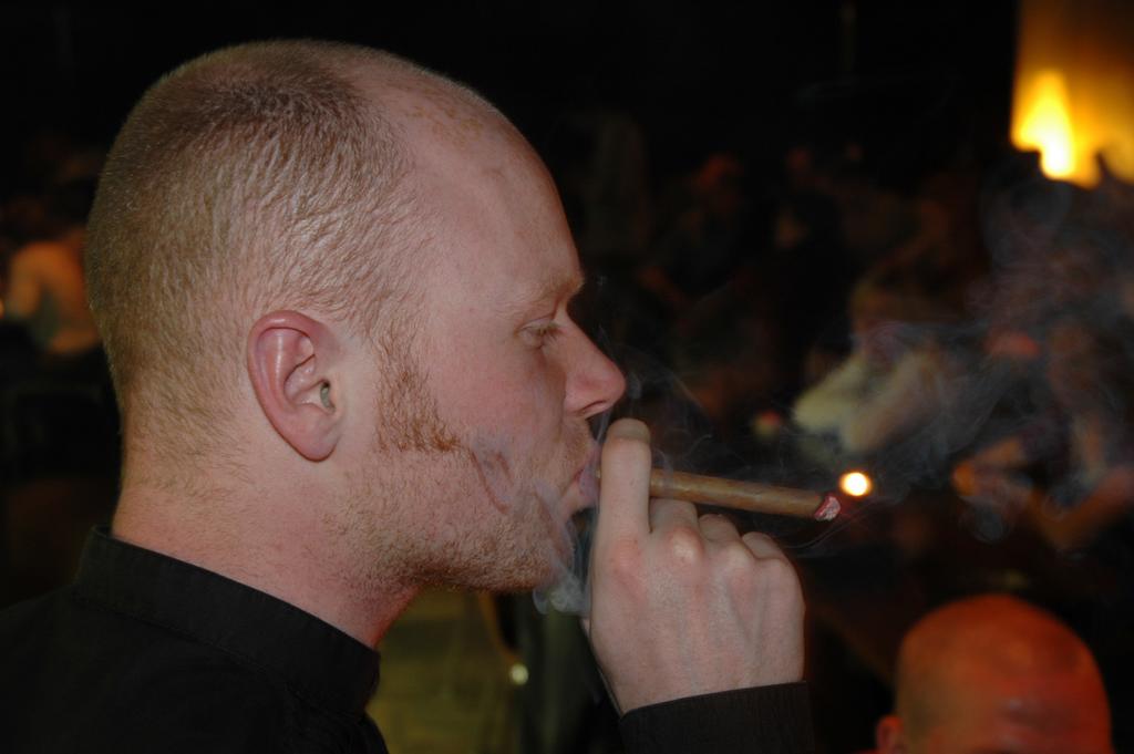A man in a black shirt smokes a cigar, exhaling smoke in a dimly lit social setting.