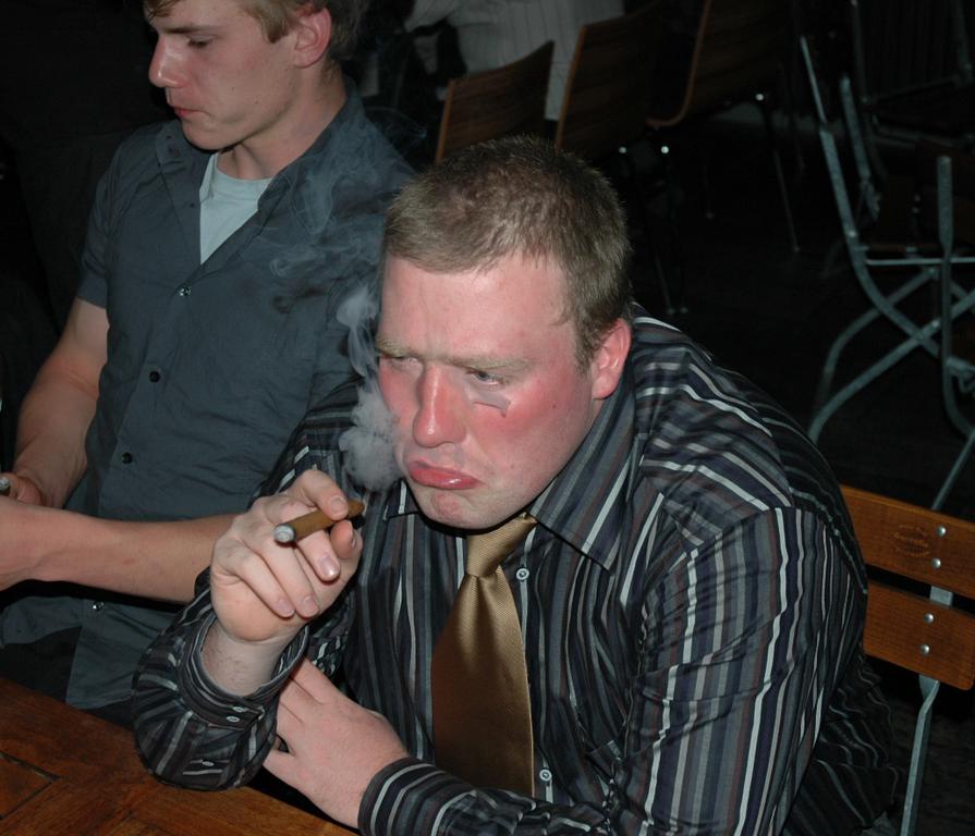 A man with a flushed face and smudged makeup smokes a cigar while sitting at a table.
