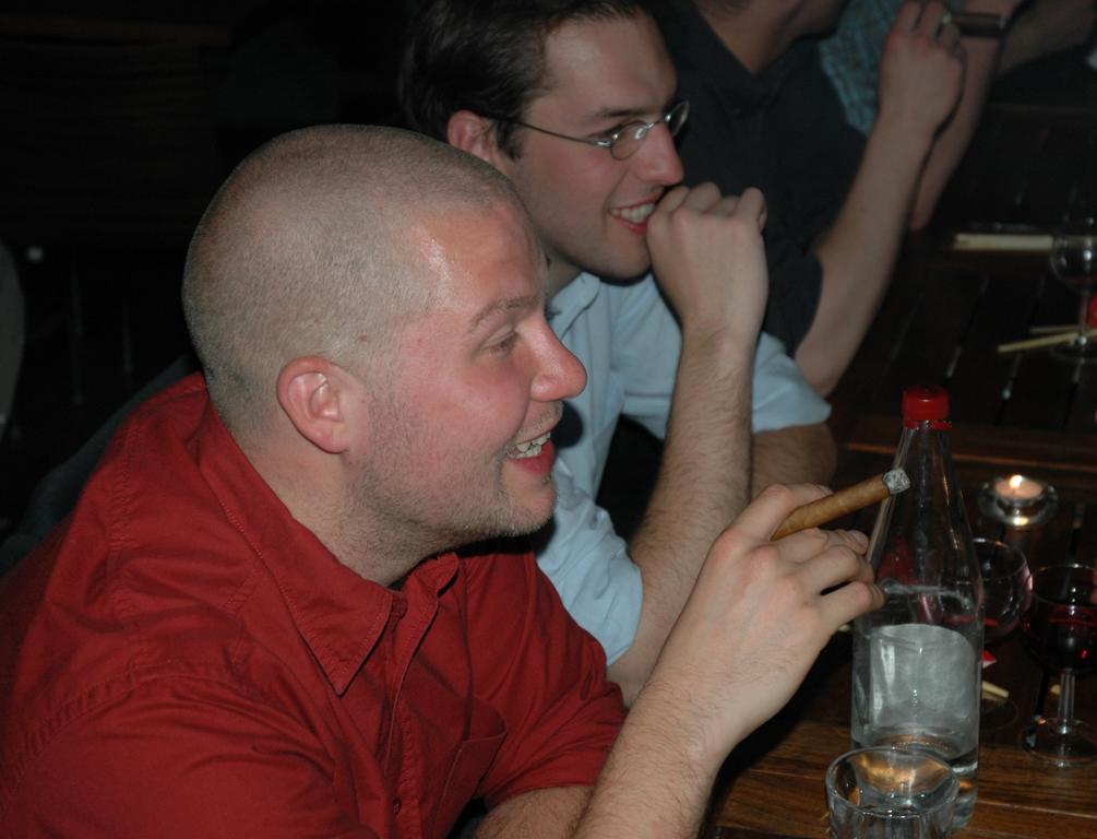 A man in a red shirt smiles while holding a cigar at a table with drinks and friends.