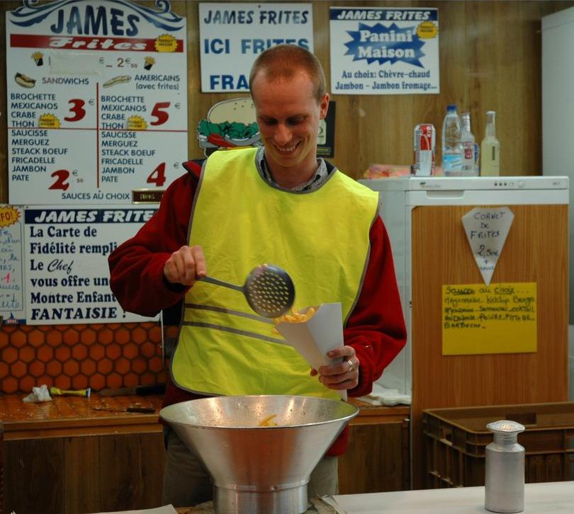 A man in a yellow vest smiles while serving fries into a paper cone at a food stand.