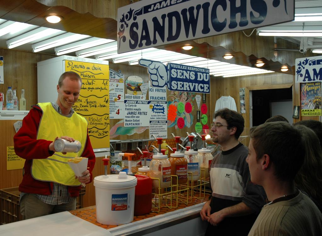 A man in a yellow vest smiles while pouring sauce on fries at a sandwich shop, with customers watching.