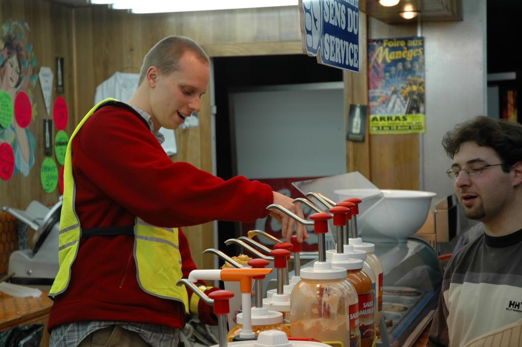 A man in a red sweater and safety vest pumps sauce onto food at a condiment station.