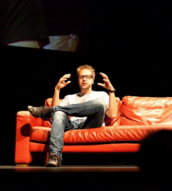 A speaker sits on a red couch, gesturing with both hands while talking during the Badcamp keynote chat.
