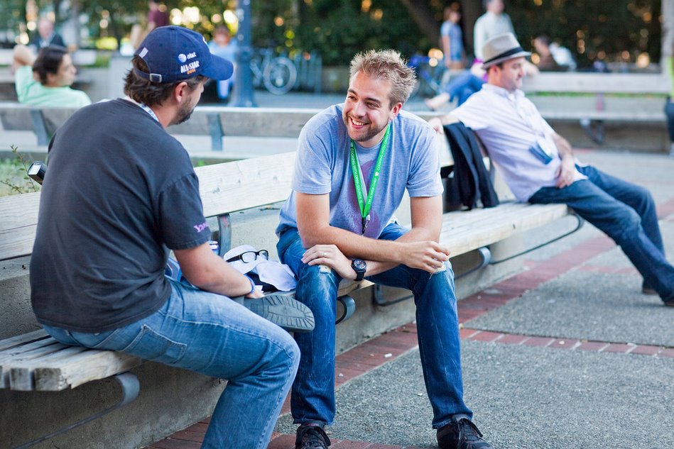 Two men sit on a bench, smiling and talking at an outdoor event.