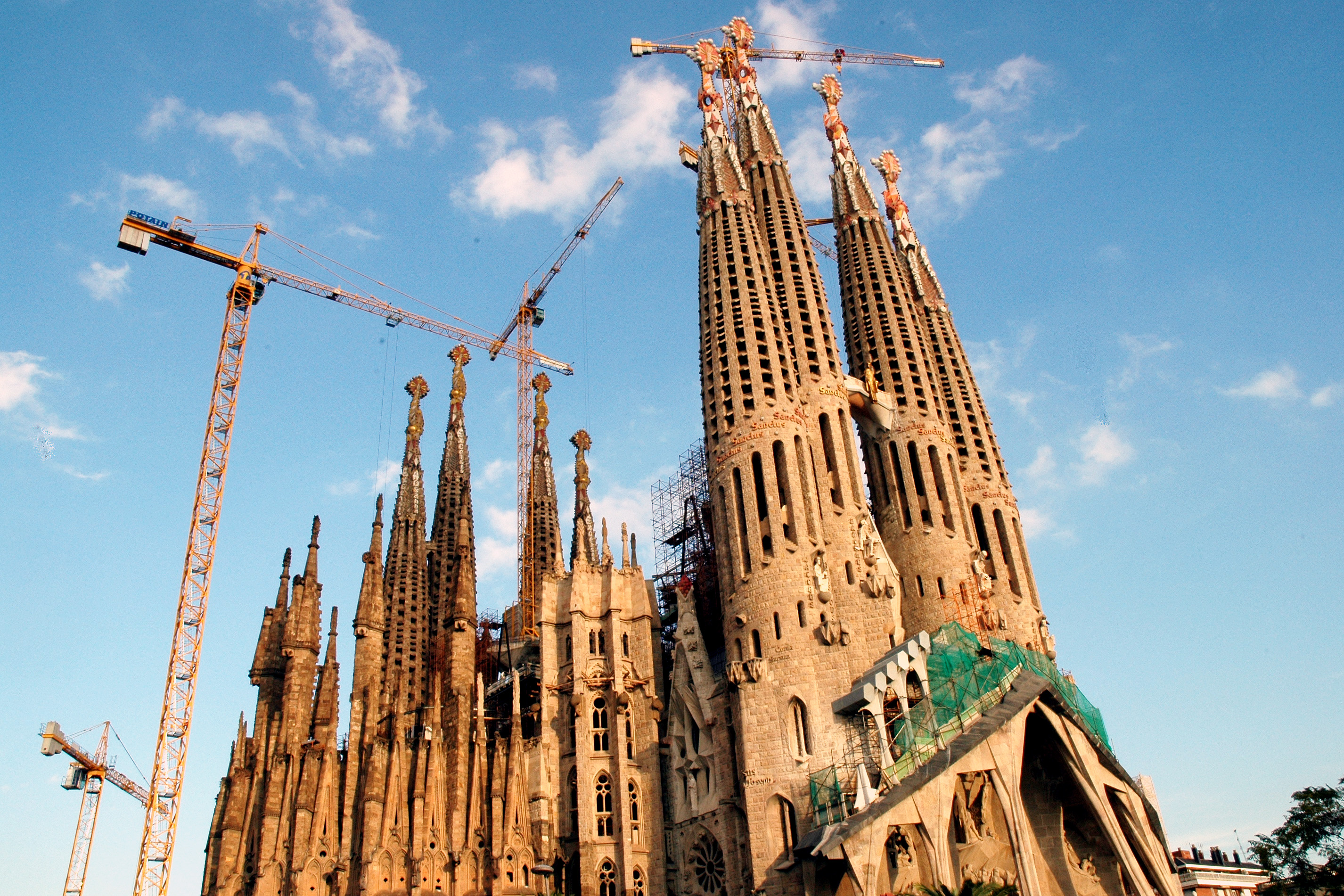 La Sagrada Familia under construction, with several cranes surrounding its tall, detailed towers against a blue sky.