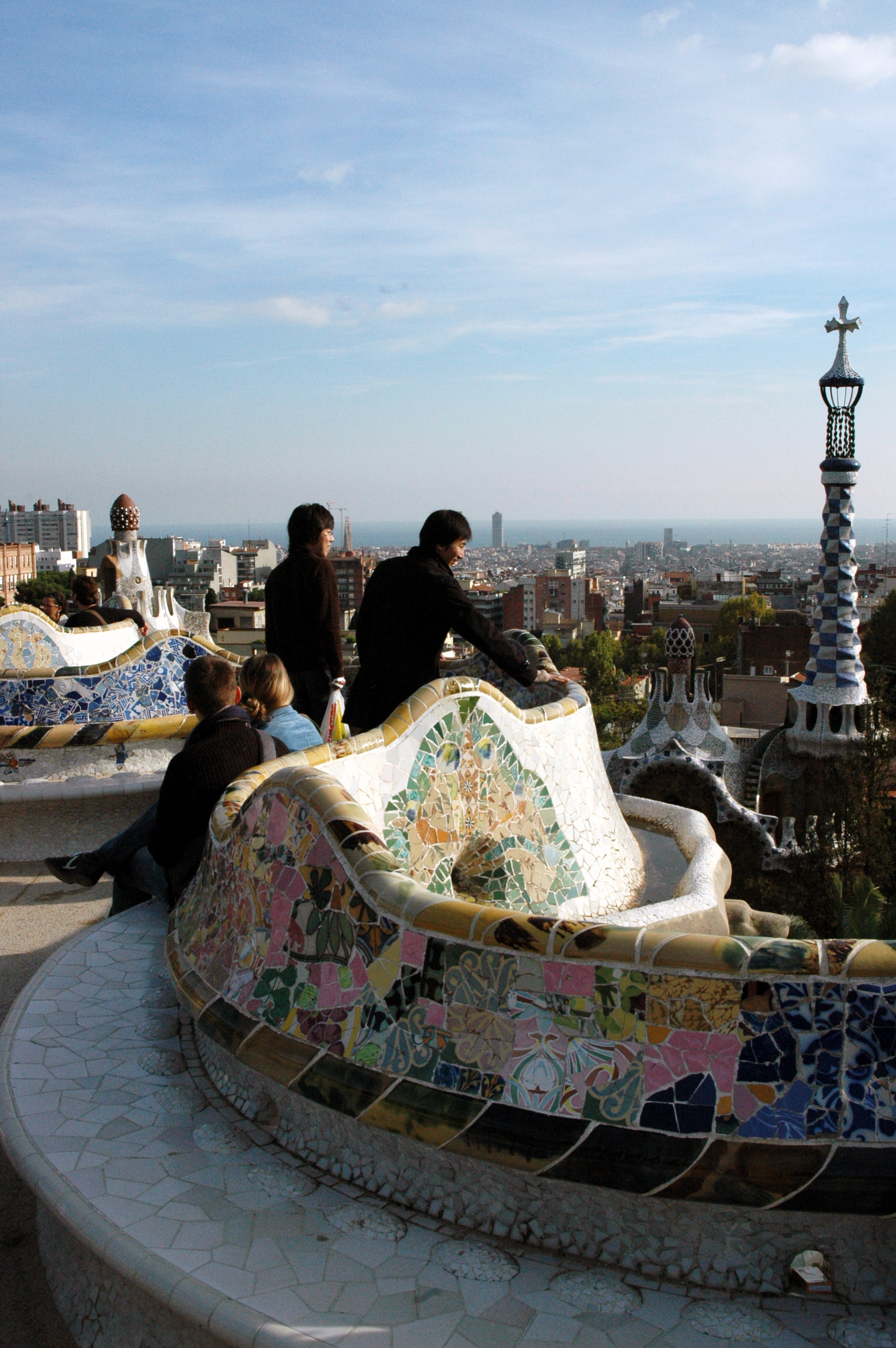 People sit and stand on colorful mosaic benches at Park Güell, overlooking the cityscape of Barcelona.