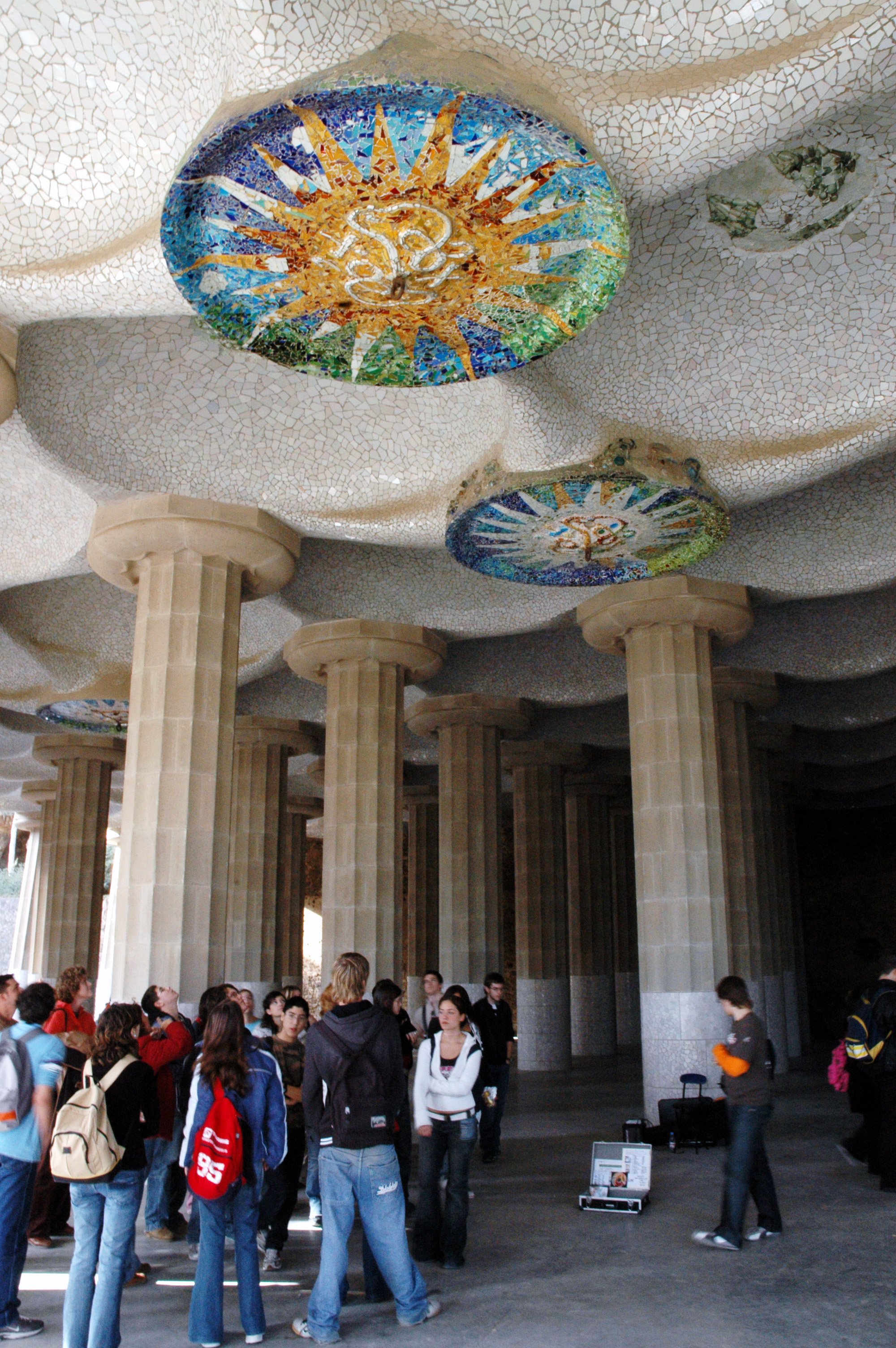 A group of people gathers under mosaic-decorated ceilings supported by stone columns in Park Güell, Barcelona.