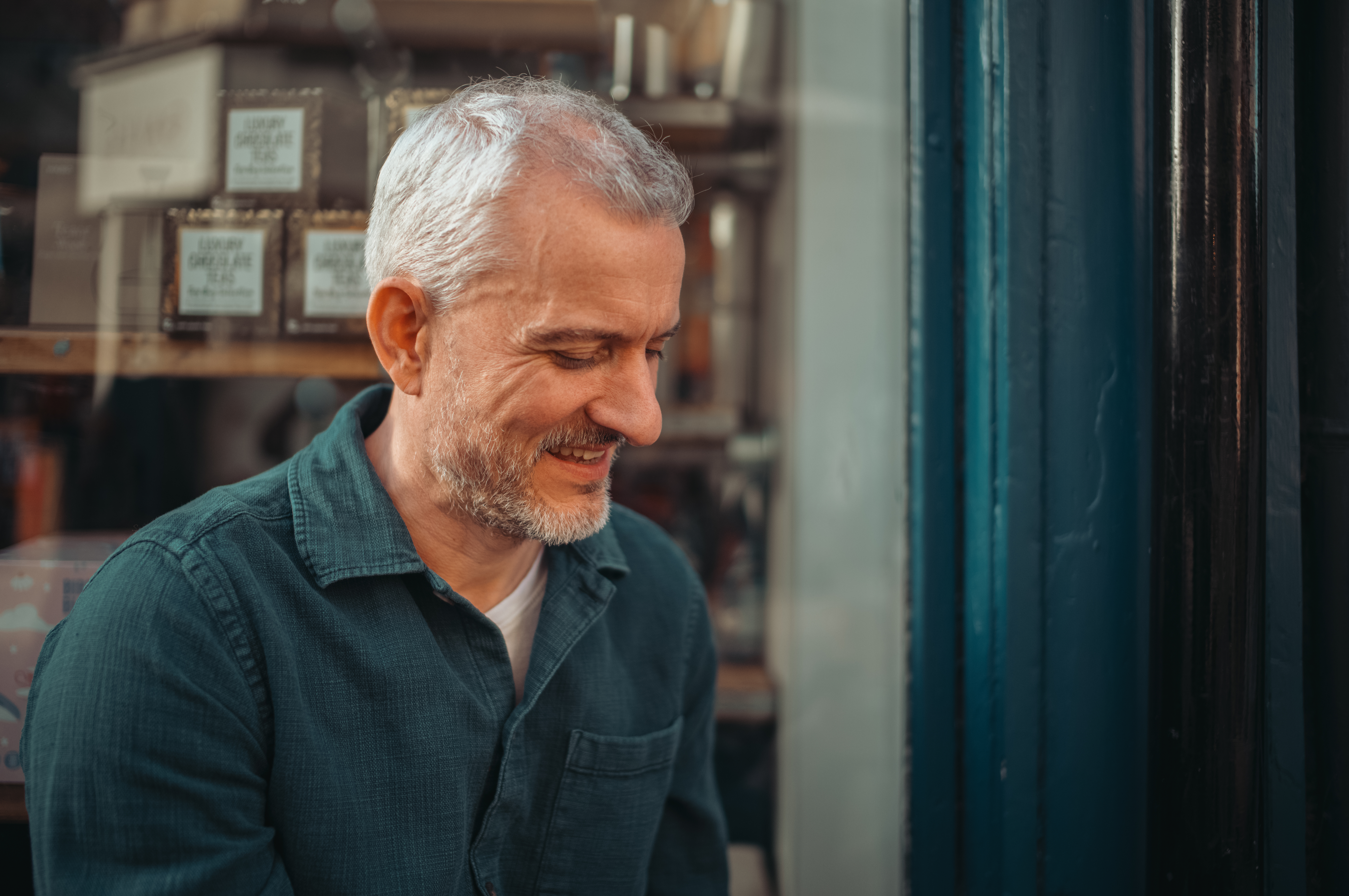 A smiling man with gray hair looking down, outside a shop window.