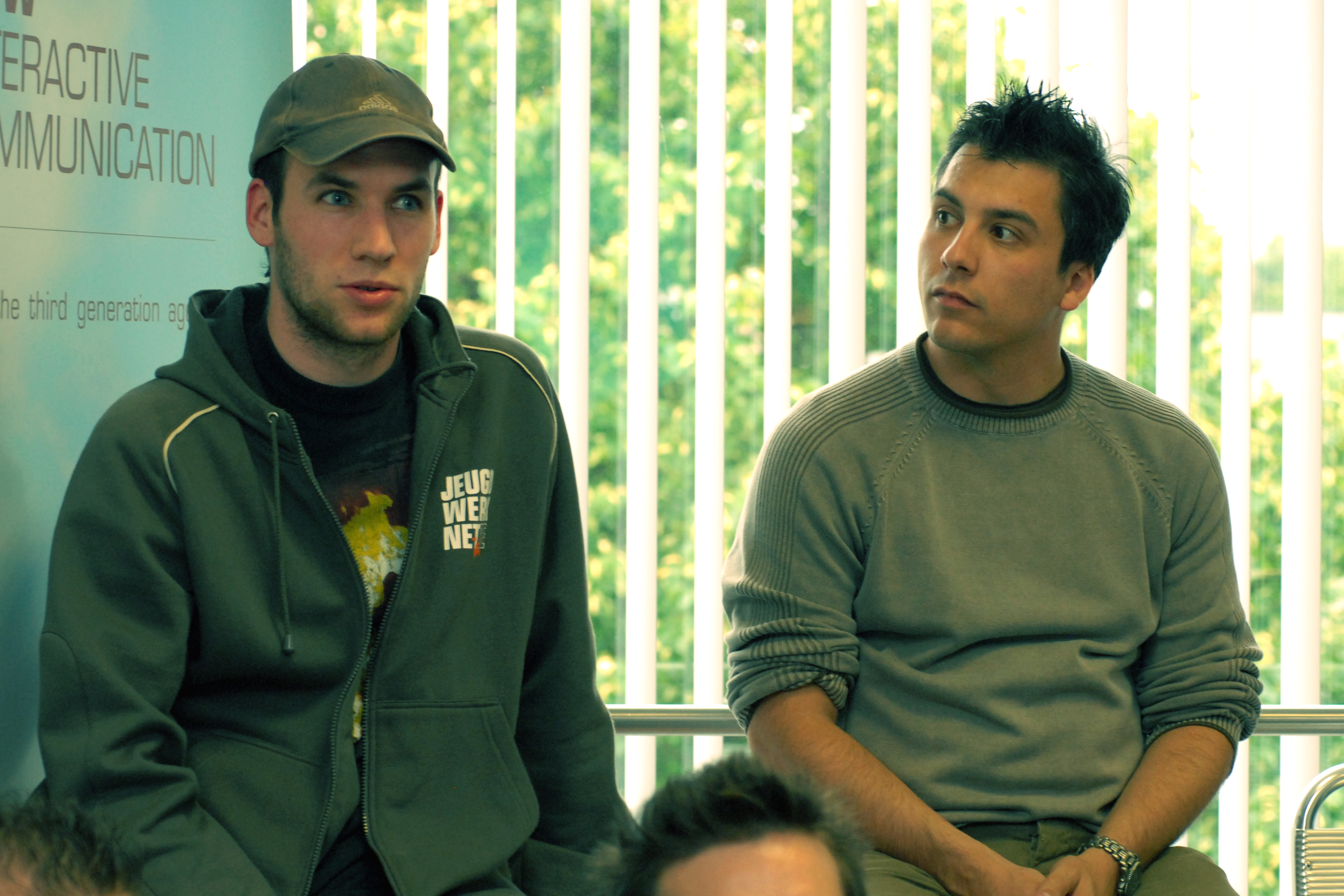 Two men sit indoors near a window, one speaking while the other listens attentively.