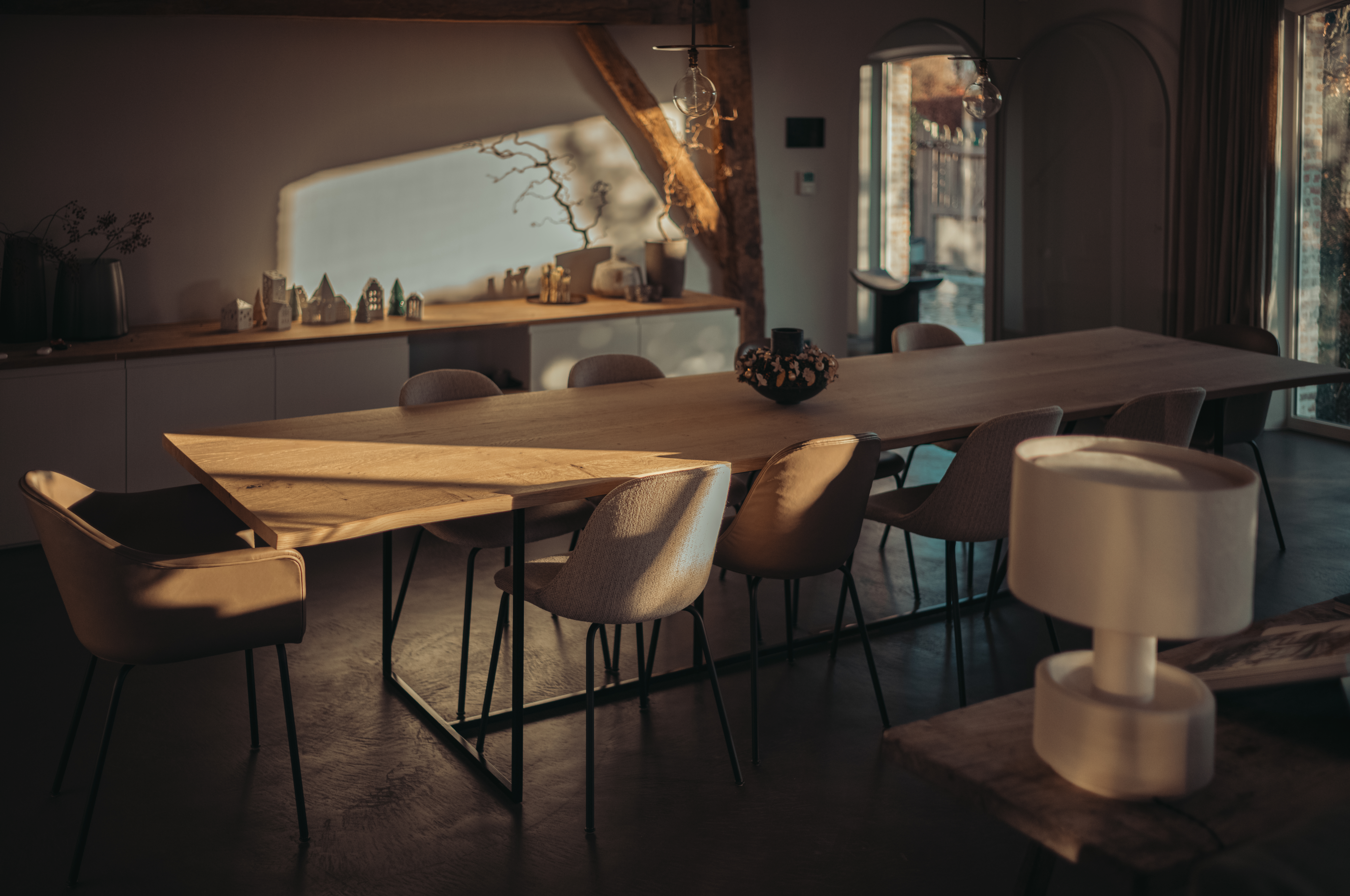 A warmly lit dining room with a long wooden table, upholstered chairs, exposed wooden beams, and sunlight casting shadows across the space.