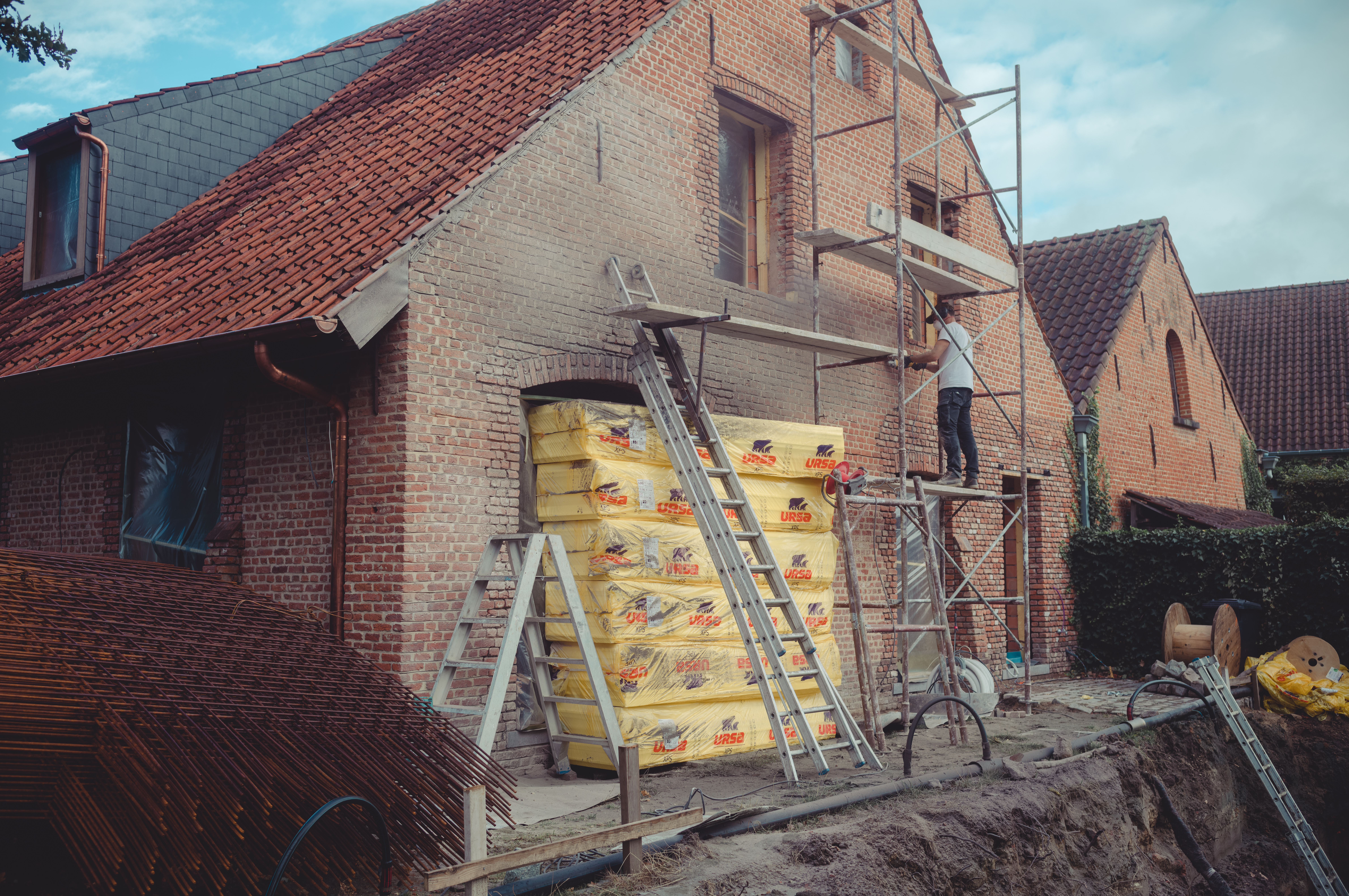 A worker on scaffolding removes old mortar from a brick wall during renovation.