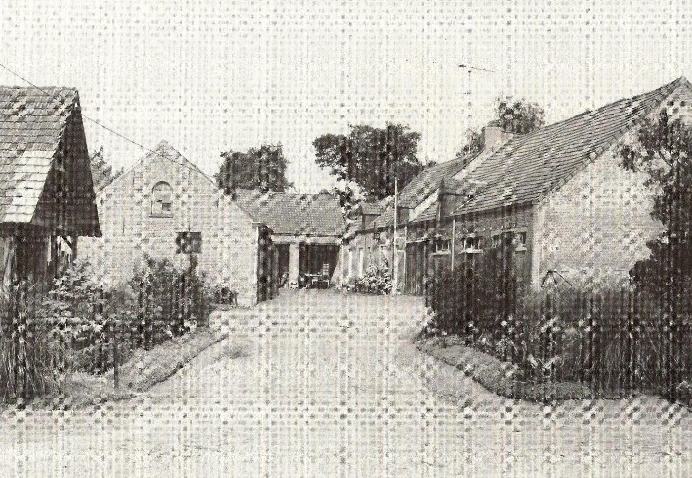 Black-and-white photo of an old brick barn and farmhouse with a courtyard, surrounded by plants and trees.