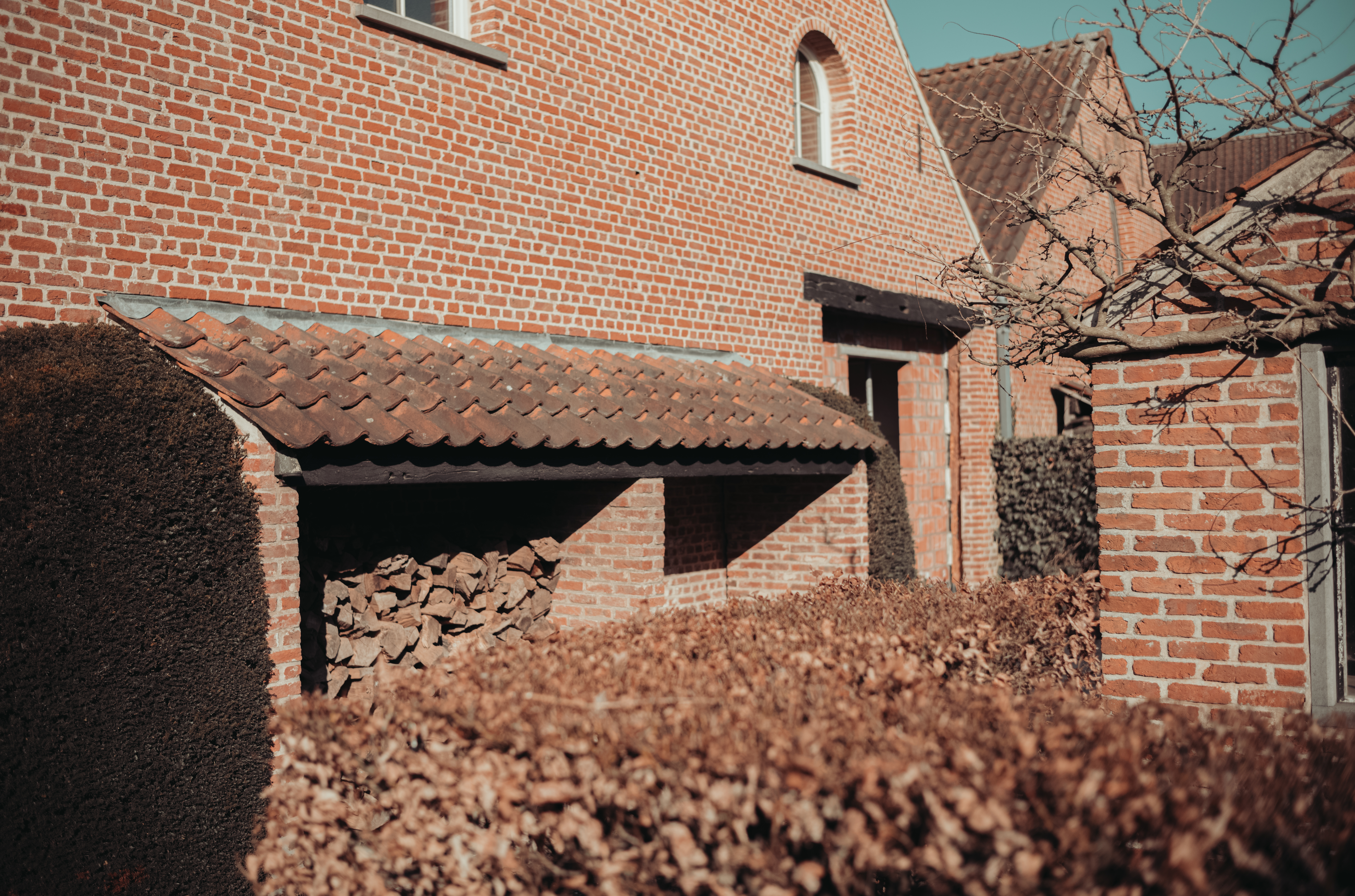 A brick house with a wood storage area under a tiled roof, partially hidden by hedges.