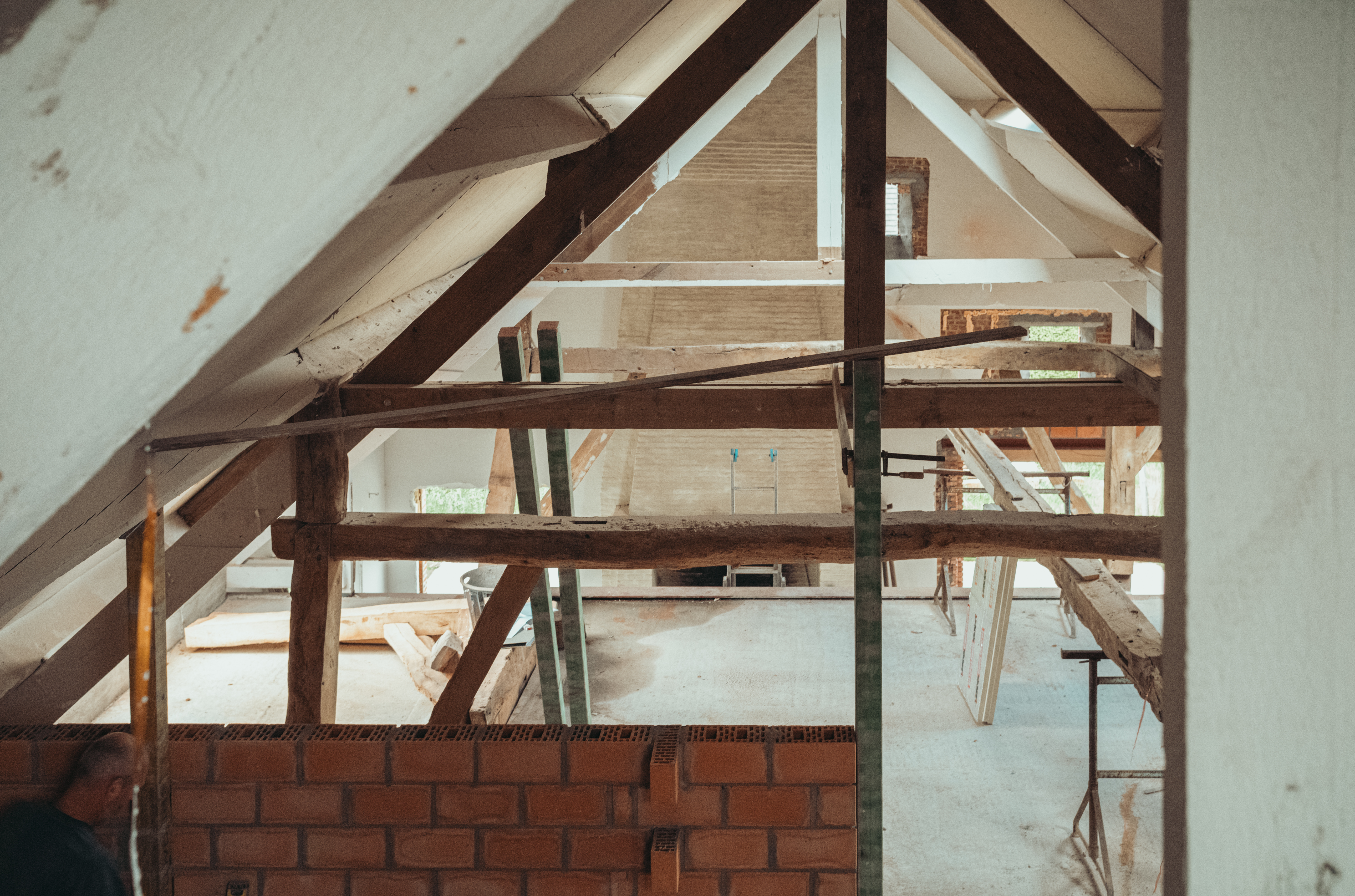 Partially constructed barn roof with exposed wooden beams, brick wall, and construction materials scattered throughout the space.