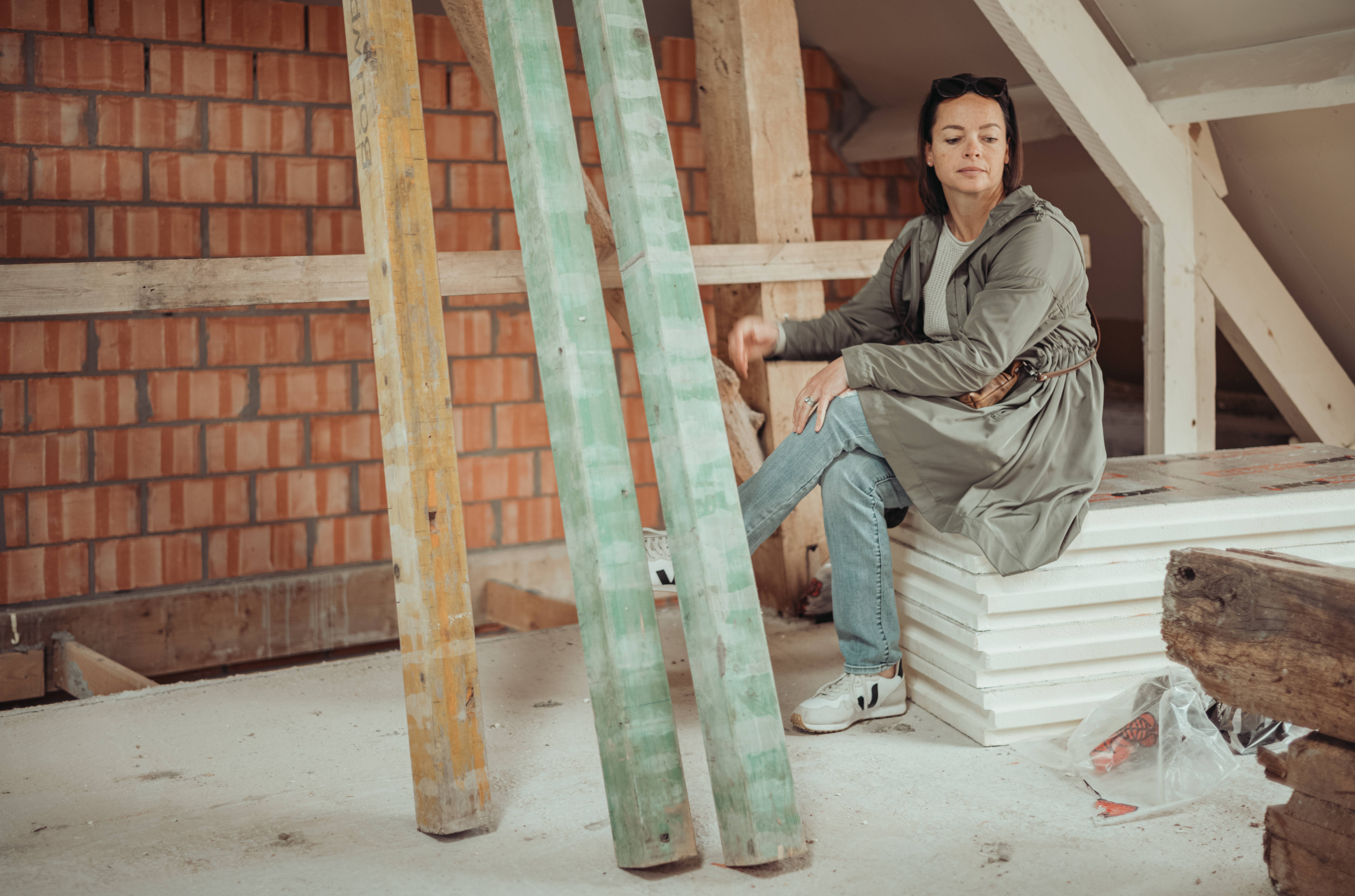 A woman sits on stacked insulation sheets in a partially constructed space, wearing a green coat and jeans.