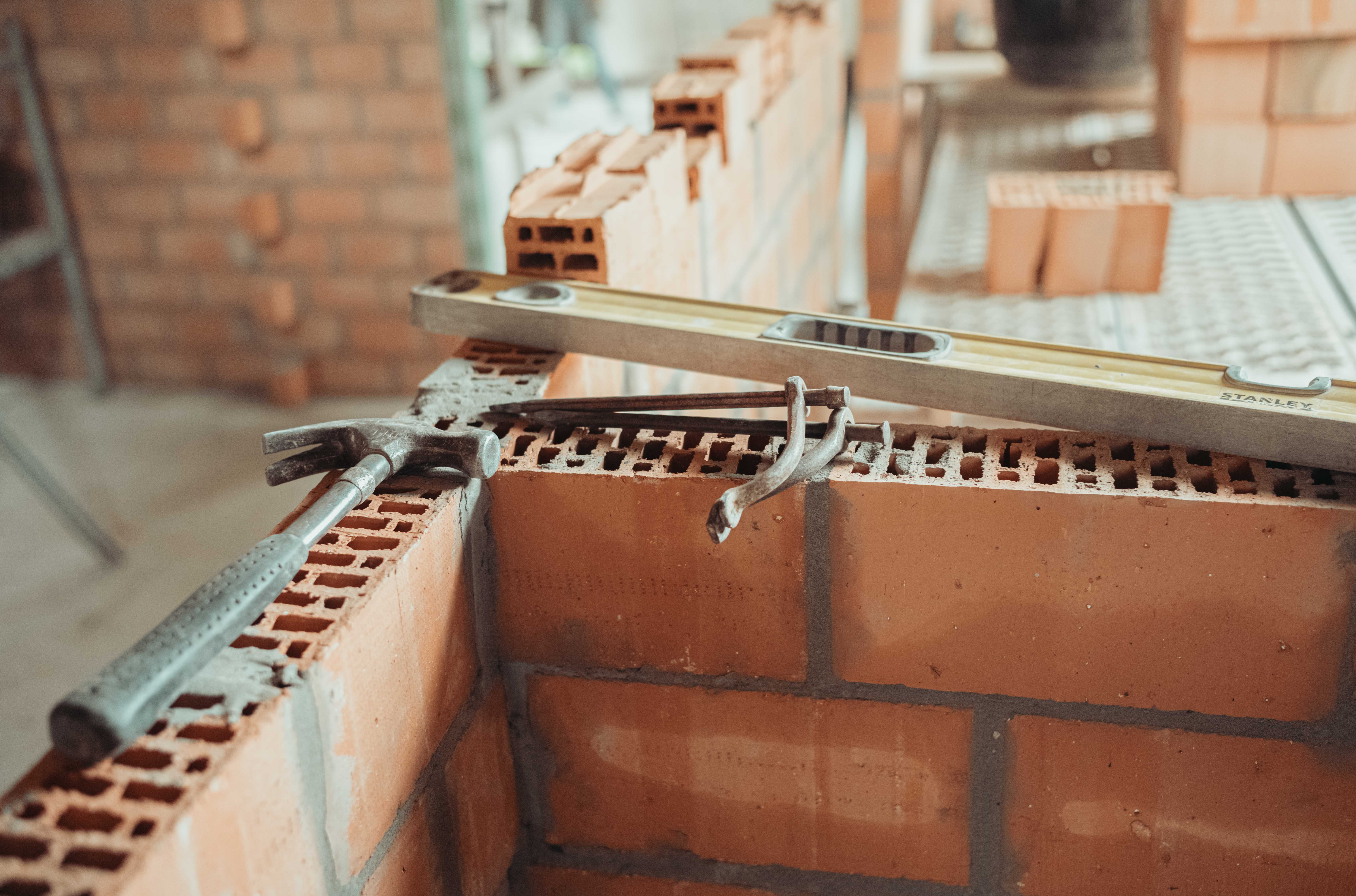 A hammer, a brick trowel, and a level rest on a partially built brick wall at a construction site.