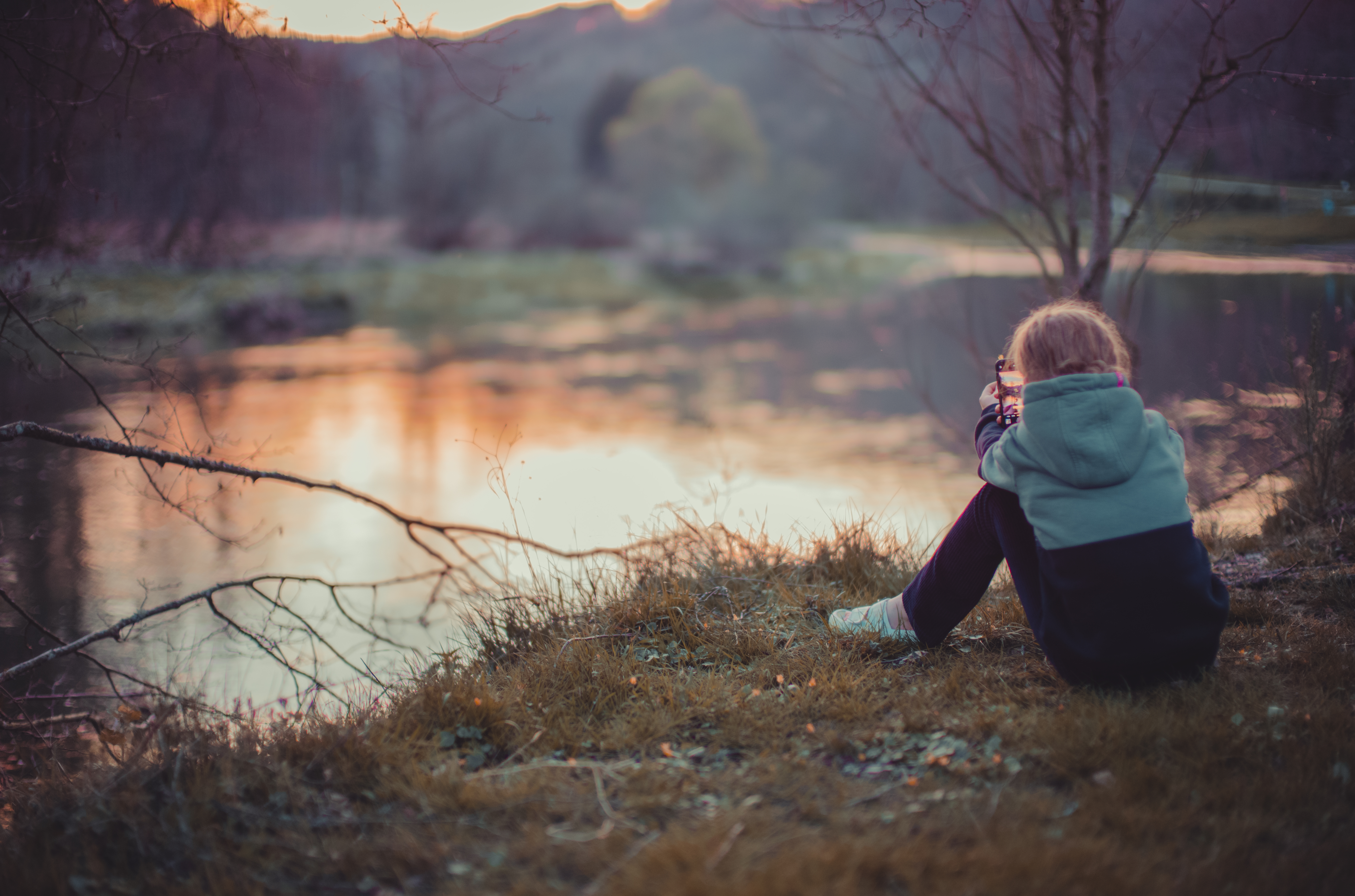 A person in a hoodie sits by the water, aiming a small object with both hands.