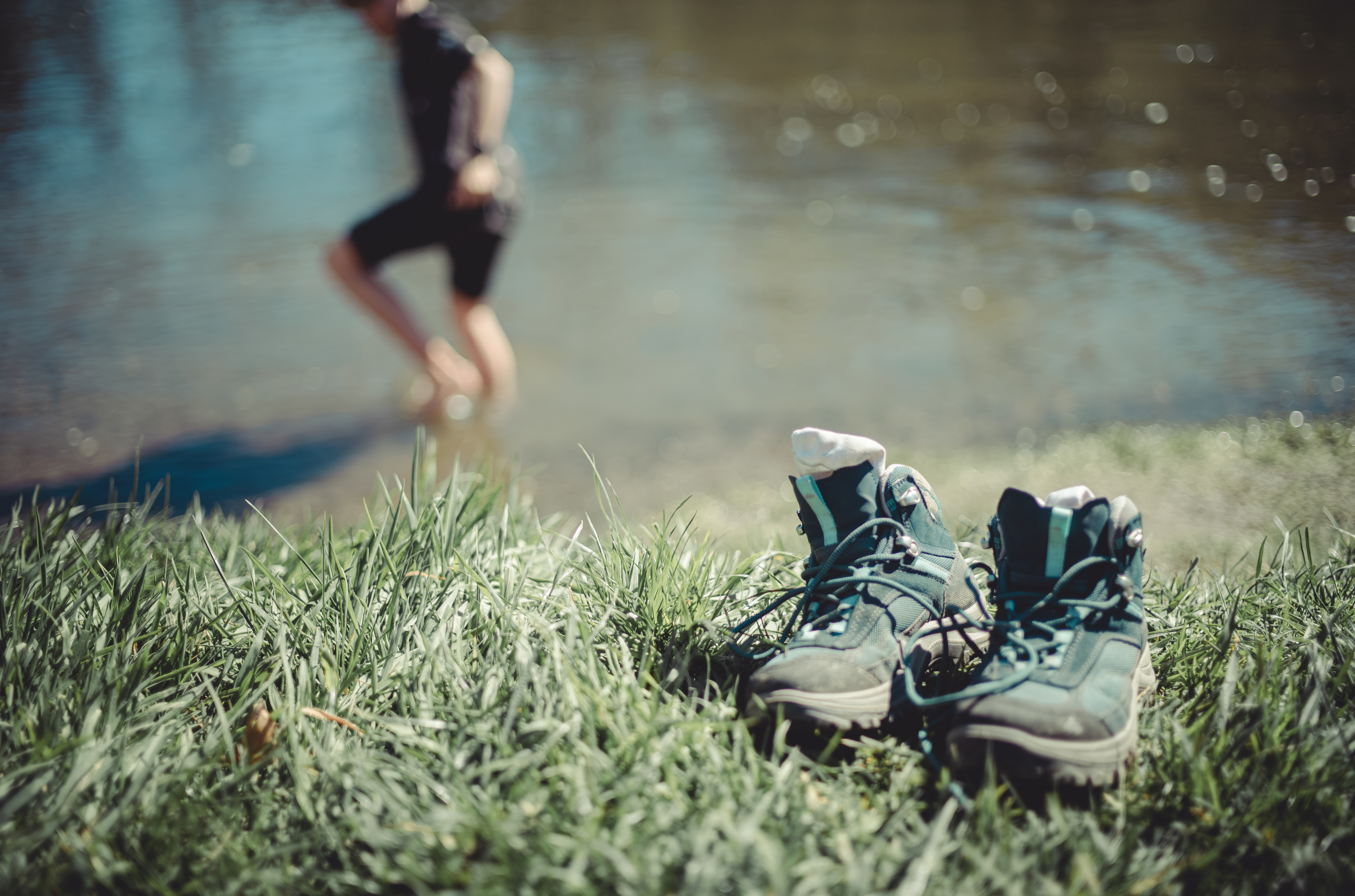 A pair of hiking boots on grass, with a person wading in the water in the background.