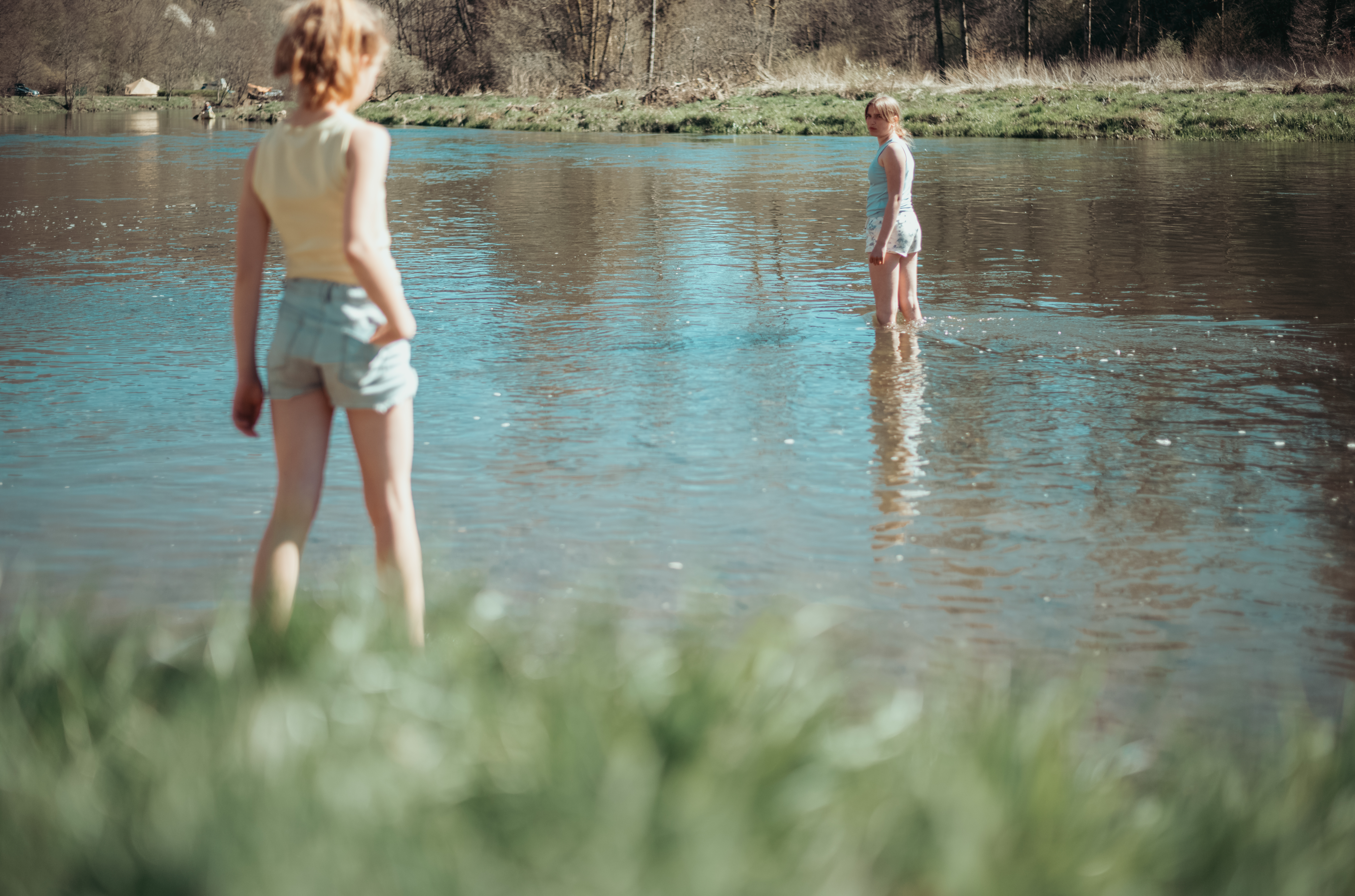 Two children stand in a shallow river, one in the water and the other near the edge, looking around.