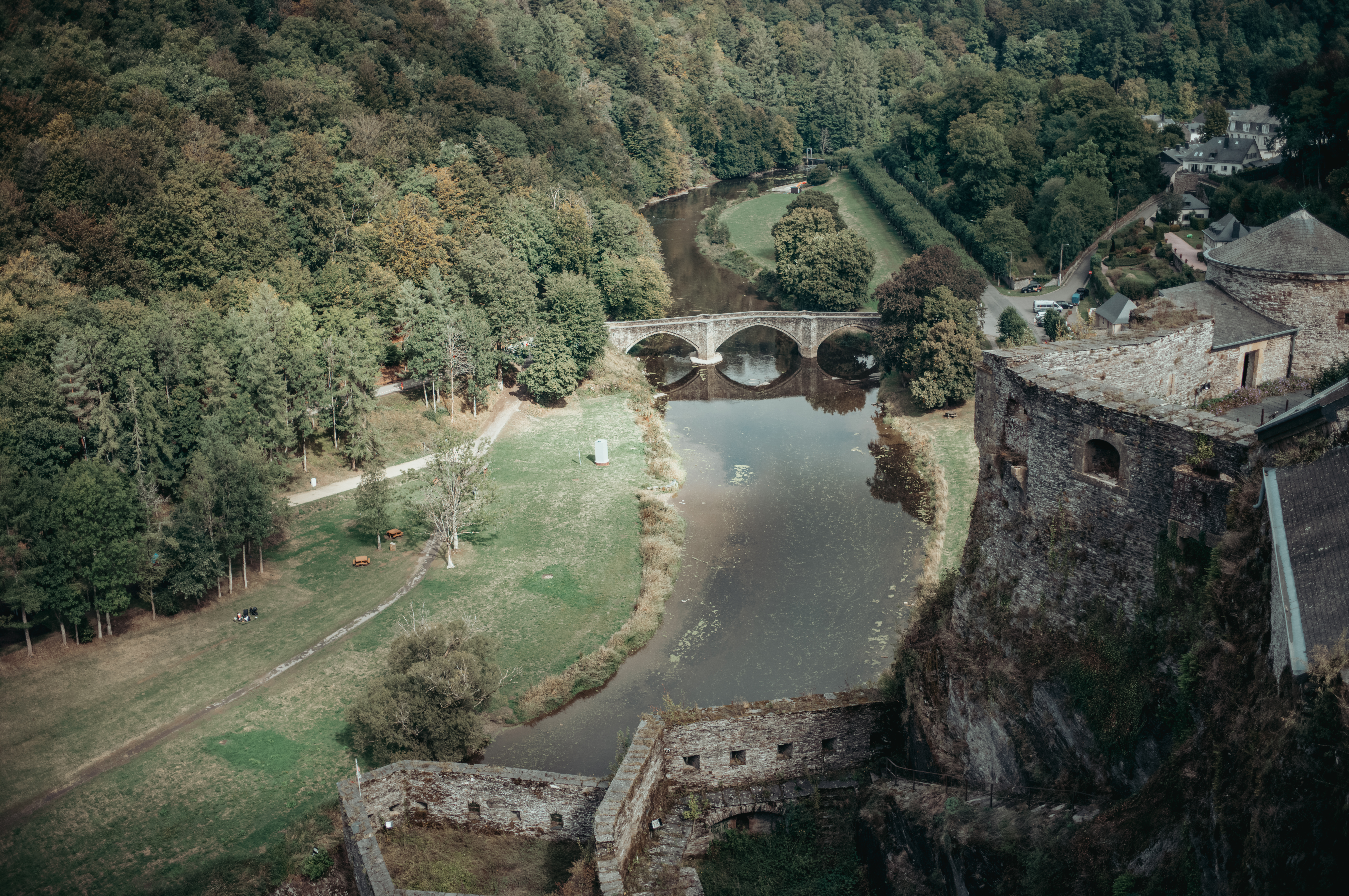 Castle walls rising above a river with a medieval bridge and green banks..